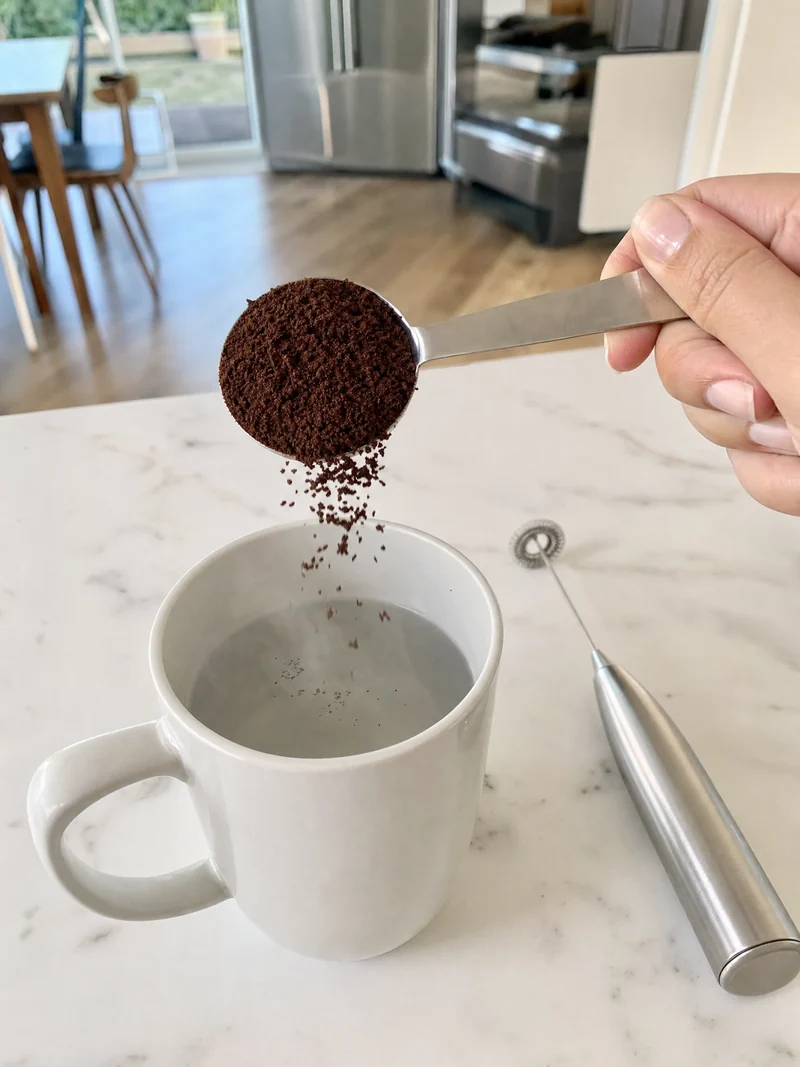 A tablespoon of dark coffee powder being tipped into a mug of hot water beside a frother